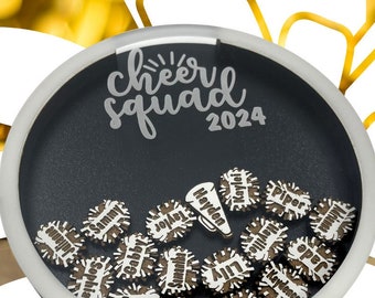 May include: A clear round container with a black background and white lettering that says "cheer squad 2024". Inside the container are small wooden circles with names written on them, including "Hayden", "Lily", "Ava", "Piper", "Sophia", "Gabby", "Ellie", "Jolie", "Summer", "Trinity", "Katie", "Jenna", and "Jamie".