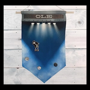 May include: A blue and white fabric banner with the word "OLE" printed on it. The banner has a distressed look and is hanging from a wooden dowel. There are several small pins attached to the banner.