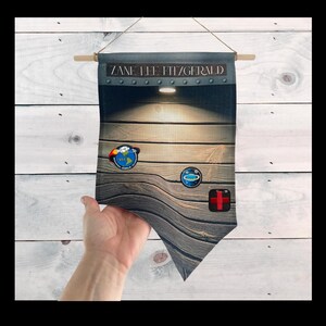 May include: A wooden wall banner with the name "Zane Lee Fitzgerald" printed on it. The banner has a light shining on it and features three patches: a blue and white Earth patch, a blue and white Saturn patch, and a red cross patch.