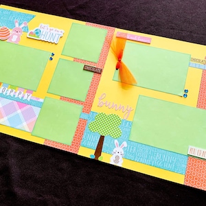 以下が含まれることがあります： 緑色の写真スペースがある、黄色のイースターをテーマにしたスクラップブックページ。装飾要素には、ピンクのウサギ、イースターエッグ、リボン、「HOPPY」、「bunny」、「Chocolate Bunnies」の文字が含まれています。