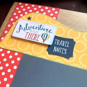 May include: A scrapbook page with a yellow and white sunflower pattern, a red and white polka dot pattern, and a black background. The page has a blue and white sign that says "Adventure is out there" with a hot air balloon graphic. There is also a black sign that says "Travel Notes".