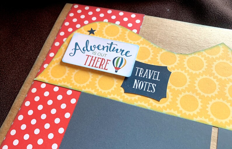 May include: A scrapbook page with a yellow and white sunflower pattern, a red and white polka dot pattern, and a black background. The page has a blue and white sign that says "Adventure is out there" with a hot air balloon graphic. There is also a black sign that says "Travel Notes".