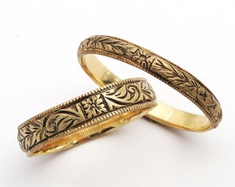 May include: Two gold wedding bands with intricate floral engravings. The bands are stacked on top of each other.