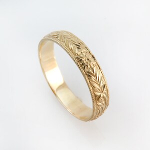 May include: A gold band ring featuring an intricate engraved pattern of repeating leaves and small star-like motifs. The edges of the ring are finished with a delicate beaded texture, providing a detailed border along the entire circumference.