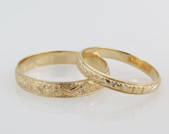 May include: Two gold wedding bands with intricate floral engravings. The bands are stacked on top of each other.