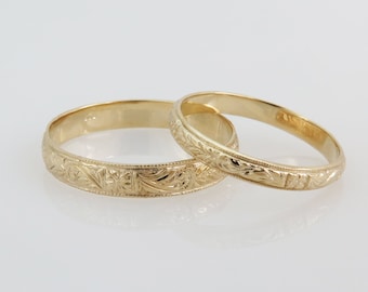 May include: Two gold wedding bands with intricate floral engravings. The bands are stacked on top of each other.