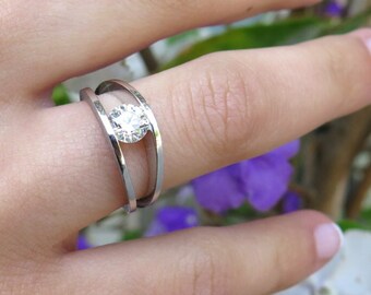 Puede incluir: Un anillo de plata con una sola piedra preciosa redonda y transparente engastada en un engaste de bisel. El anillo tiene un diseño único con dos bandas que se cruzan.