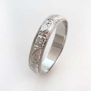 May include: A silver-toned metal ring featuring an intricate engraved floral and leaf pattern along the outer band. The edges of the ring are defined by a delicate beaded texture, providing a detailed border to the central embossed design.