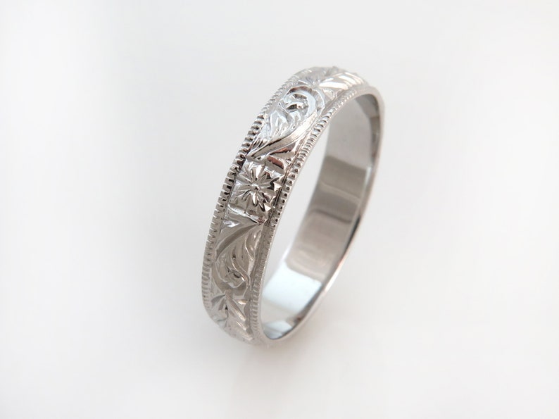May include: A silver-toned metal ring featuring an intricate engraved floral and leaf pattern along the outer band. The edges of the ring are defined by a delicate beaded texture, providing a detailed border to the central embossed design.