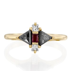 May include: A gold ring featuring a geometric design with a central rectangular red gemstone, flanked by two dark gray triangular stones and two square white gemstones. The ring has a delicate band.