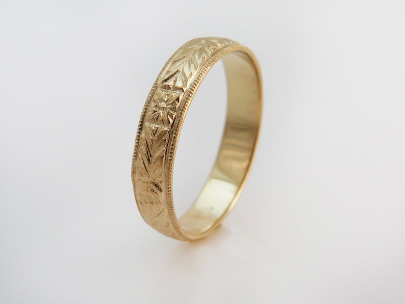 May include: A yellow gold band ring featuring a repeating engraved pattern of leaves and star-like shapes along the outer surface. The edges of the ring are defined by a fine beaded milgrain texture, and the interior of the band is smooth and polished.
