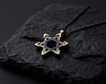 May include: A Star of David pendant necklace. The pendant features a six-pointed star design with gold-colored accents and a black center. The necklace chain is dark gray. The pendant is displayed on a dark, textured surface.