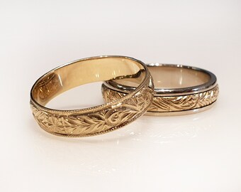 May include: Two gold wedding bands with intricate floral engravings. The band on top is a thinner band with a smooth inner surface. The band on the bottom is a thicker band with a textured inner surface.
