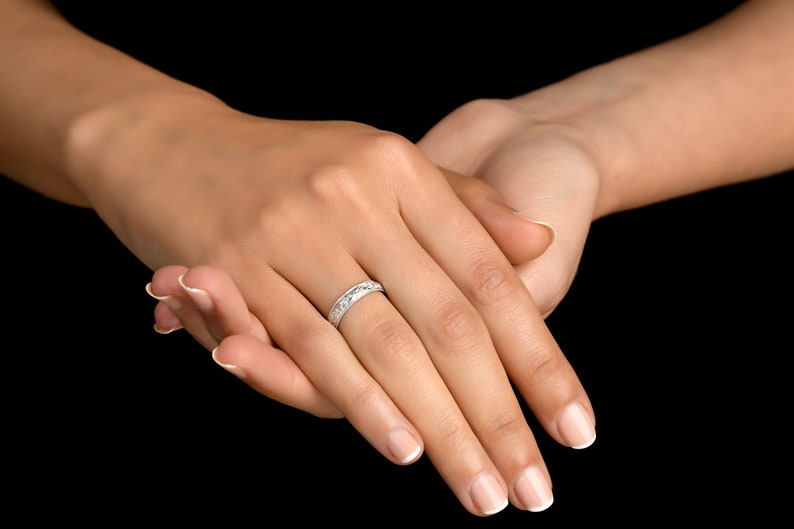 May include: A silver-toned band ring featuring a repeating engraved pattern along its outer surface. The ring has a polished metallic finish and a consistent width, encircling the finger with a detailed, textured design that catches the light.