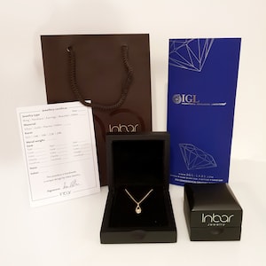 May include: A brown jewelry box with a gold necklace inside. The box is open and the necklace is visible. The necklace has a small, teardrop-shaped pendant. The box is sitting on a white surface. The image also includes a brown paper bag with the word "Inbar" printed on it, a blue card with the word "IGL" printed on it, and a smaller brown jewelry box with the word "Inbar" printed on it.