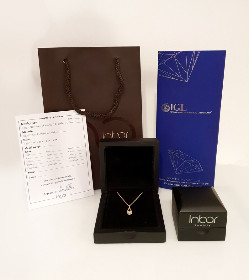 May include: A brown jewelry box with a gold necklace inside. The box is open and the necklace is visible. The necklace has a small, teardrop-shaped pendant. The box is sitting on a white surface. The image also includes a brown paper bag with the word "Inbar" printed on it, a blue card with the word "IGL" printed on it, and a smaller brown jewelry box with the word "Inbar" printed on it.