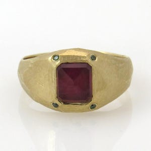 Puede incluir: Un anillo de oro con una gran piedra preciosa roja de talla esmeralda rodeada de cuatro piedras preciosas verdes más pequeñas.