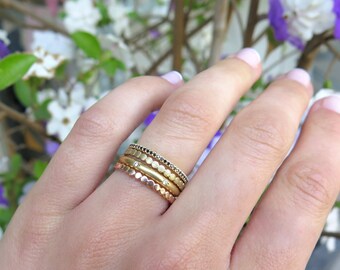 May include: A set of five gold stacking rings. The rings are different widths and have various textures, including hammered, smooth, and beaded. The rings are worn on a finger.