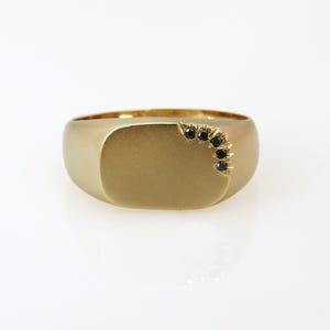 May include: A gold signet ring with a matte finish featuring a smooth, flat rectangular face. A curved row of five small, round, black gemstones is set into the upper right corner of the ring face, creating a subtle, dark accent against the gold.
