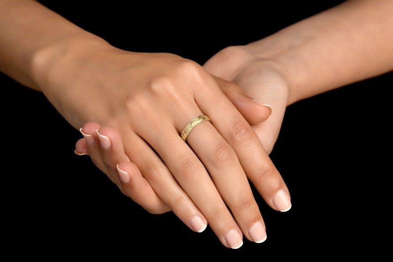 May include: A yellow gold band ring featuring a delicate, repeating engraved floral pattern across its entire outer surface. The ring has a polished finish and a slim, rounded profile, showcasing intricate botanical details etched into the metal.
