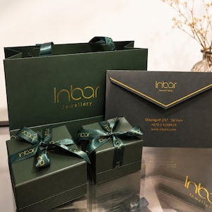 May include: A dark green shopping bag, two dark green square gift boxes tied with matching branded ribbons, and a dark grey envelope. Each item features the gold Inbar Jewellery logo. The envelope includes the address Dizengoff 237, Tel Aviv.