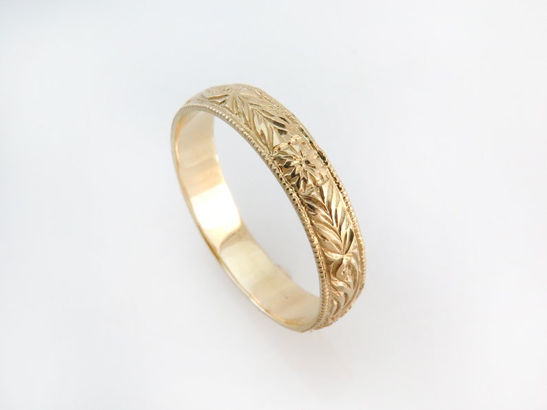 May include: A gold band ring featuring an intricate engraved pattern of repeating leaves and small star-like motifs. The edges of the ring are finished with a delicate beaded texture, providing a detailed border along the entire circumference.