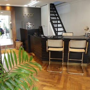 May include: A commercial interior space featuring a black reception desk with two matching bar stools with cane backrests and seats, gold-toned metal frames, and a dark gray wall displaying the gold text Inbar. A black staircase is visible in the back.