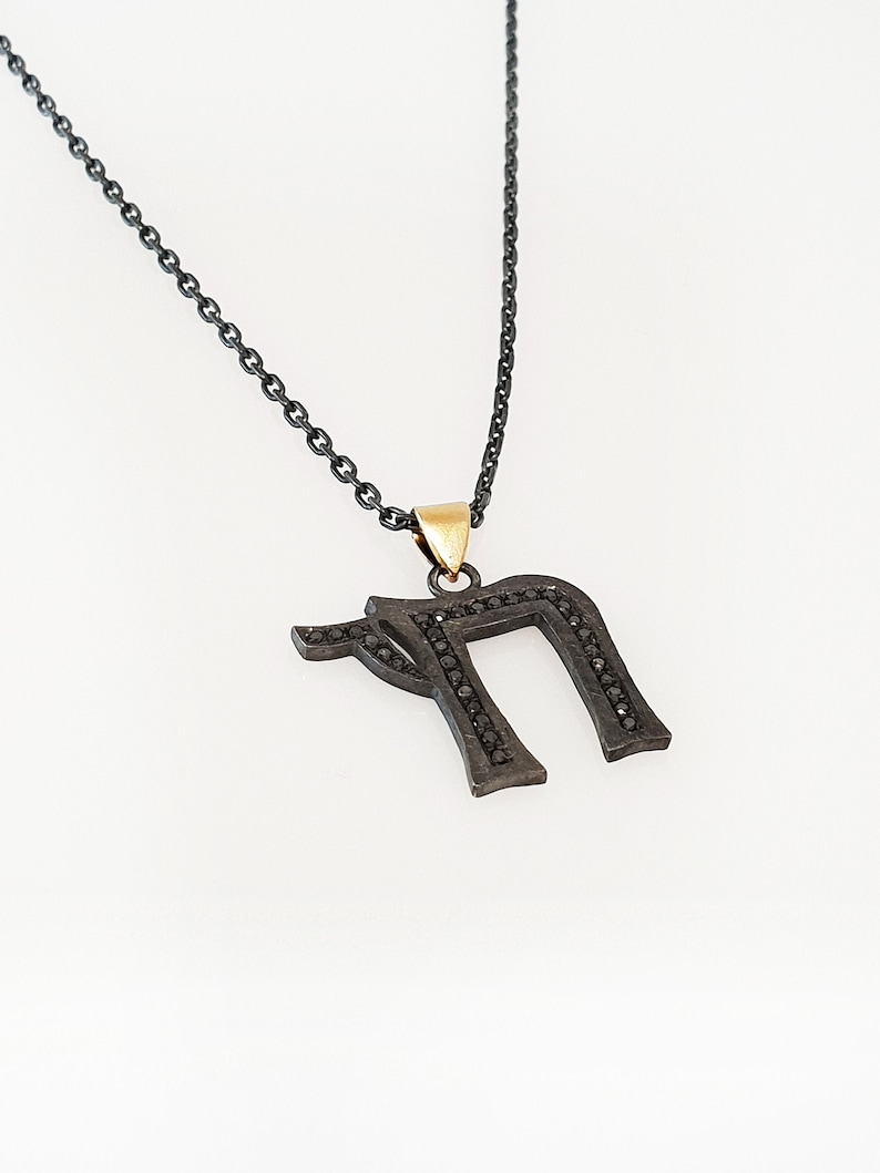 May include: A black chain necklace with a black metal pendant in the shape of the Hebrew letter 'nun' with black diamonds. The pendant has a gold bail.