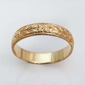 May include: A gold band ring featuring an intricate engraved floral pattern along the outer surface. The ring has a polished finish with a beaded milgrain edge detail running along both sides of the central decorative band design.