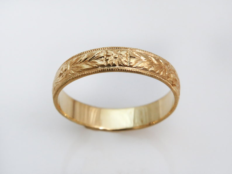 May include: A gold band ring featuring an intricate engraved floral pattern along the outer surface. The ring has a polished finish with a beaded milgrain edge detail running along both sides of the central decorative band design.