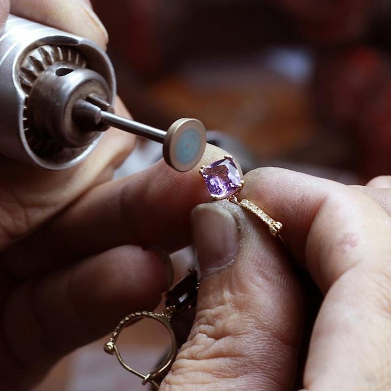 May include: A gold ring featuring a square-cut light purple gemstone being polished by a small rotary tool. The ring band has a textured surface and is held in place while the metal tool with a circular buffing head works on the setting.