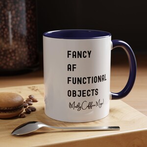 May include: White ceramic mug with a navy blue interior and handle. The mug features the text "FANCY AF FUNCTIONAL OBJECTS Mostly Coffee Mugs" in black. A silver spoon and a macaron are next to the mug.