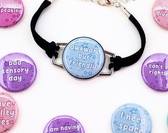 Könnte beinhalten: Ein schwarzes Armband mit einem hellblauen Anhänger mit der Aufschrift "awkward but friendly". Um das Armband herum befinden sich mehrere bunte Ansteckbuttons mit verschiedenen Phrasen, darunter "I have ADHD" und "I am having a panic attack".