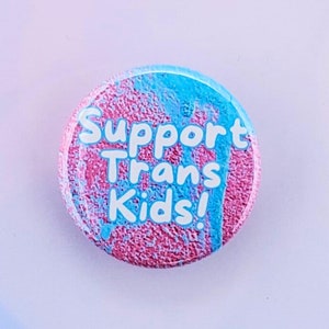 May include: A pink and blue button with the text "Support Trans Kids!" in white lettering.