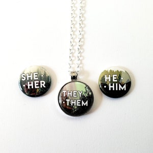 以下が含まれることがあります： 森の背景と白い文字が書かれた丸いピンが3つ。ピンには「彼女/彼女」、「彼/彼」、「彼ら/彼ら」と書かれています。「彼ら/彼ら」のピンは銀色のチェーンに繋がっています。