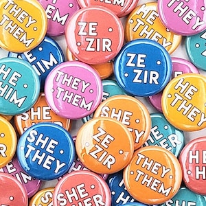 May include: A collection of colorful round buttons with different pronoun sets printed on them. The buttons include "She/Her", "He/Him", "They/Them", and "Ze/Zir".