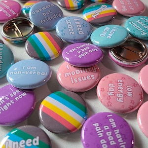 May include: A collection of round pinback buttons featuring various colors and text including "awkward but friendly," "I need space," "I have ADHD," "I am having a bad pain day," "mobility issues," "low energy day," and "I am non-verbal."