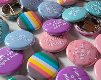 May include: A collection of round pinback buttons featuring various colors and text including "awkward but friendly," "I need space," "I have ADHD," "I am having a bad pain day," "mobility issues," "low energy day," and "I am non-verbal."