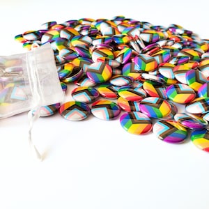 Puede incluir: Una bolsa de organza blanca llena de botones redondos de colores. Los botones presentan un patrón de chevron arcoíris con una franja marrón.