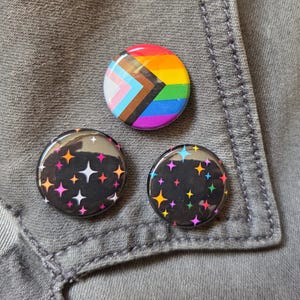 Peut inclure: Trois badges ronds. Un badge présente le drapeau Progress Pride avec des rayures arc-en-ciel horizontales et un chevron blanc, rose, bleu clair, marron et noir. Deux badges présentent un fond noir avec des étoiles colorées.