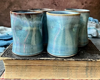 Könnte beinhalten: Vier blaue Keramikbecher mit hellblauem Überzug und braunem Rand. Die Becher stehen auf einem alten, abgenutzten Buch.
