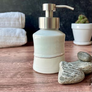 Può includere: Dispenser di sapone in ceramica bicolore con pompa in metallo spazzolato. La parte superiore è verde chiaro e la base è color crema. Accanto c'è una pila di asciugamani bianchi e un piccolo cactus in vaso.