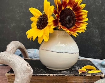 May include: A round, cream-colored ceramic vase with a smooth, glossy finish holds two sunflowers. One sunflower is bright yellow, while the other features deep red and orange petals. The vase sits atop a thick, weathered, black-covered vintage book.