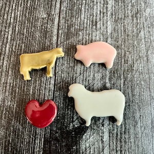 Puede incluir: Cuatro figuritas de animales de cerámica sobre un fondo de madera. Una vaca dorada, un cerdo rosa, una oveja blanca y un corazón rojo.