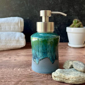 May include: A ceramic soap dispenser with a brushed metal pump. The bottle has a teal and green glaze over a grey base. The dispenser is on a wooden surface with white towels and a small cactus in the background.