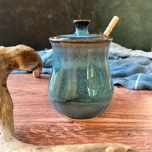 May include: A ceramic honey pot with a glossy, variegated blue and teal glaze. The pot features a matching lid with a central knob and a small opening for a wooden honey dipper, which is inserted into the top. The vessel has a rounded body.