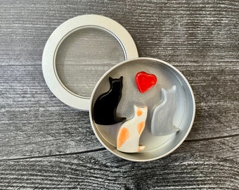 May include: A silver tin with a clear lid, containing three cat figurines and a red heart. The cats are black, white with orange spots, and gray. The tin is on a dark wood-grain surface.