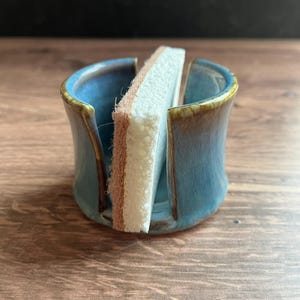以下が含まれることがあります： 光沢のある青い釉薬と茶色の縁取りが施された円筒形のセラミック製スポンジホルダー。ホルダーの両側には垂直の切り込みがあり、茶色の研磨層が付いた白い長方形のスポンジが収められています。