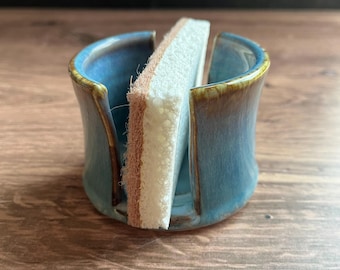 May include: A cylindrical ceramic sponge holder with a glossy blue glaze and brown rim accents. The holder features two vertical cutouts on opposite sides and contains a rectangular white sponge with a textured brown scouring layer.