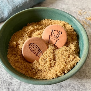 Puede incluir: Dos botes de azúcar de cerámica de color terracota con forma de cactus en un cuenco verde lleno de azúcar moreno.